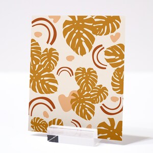 May include: A card with a white background and a pattern of brown monstera leaves and peach-colored abstract shapes. The card is on a white surface.