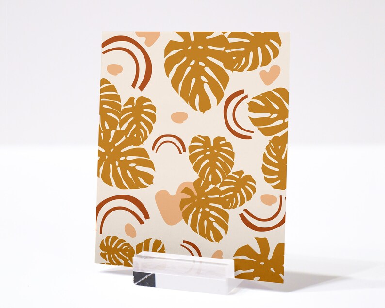 May include: A card with a white background and a pattern of brown monstera leaves and peach-colored abstract shapes. The card is on a white surface.