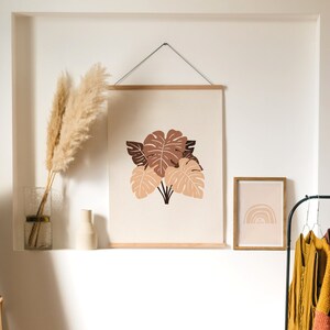 May include: A framed print with a minimalist design of brown and beige leaves on a cream background. A smaller framed print with a rainbow design is on the right. A vase with dried pampas grass is on a shelf below the prints.