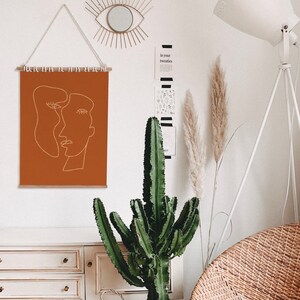 May include: A brown wall tapestry with a white line drawing of two abstract faces. The tapestry is hanging on a white wall with a decorative mirror above it. A large green cactus plant is in front of a white dresser with three drawers. A white floor lamp with a white shade is in the background.