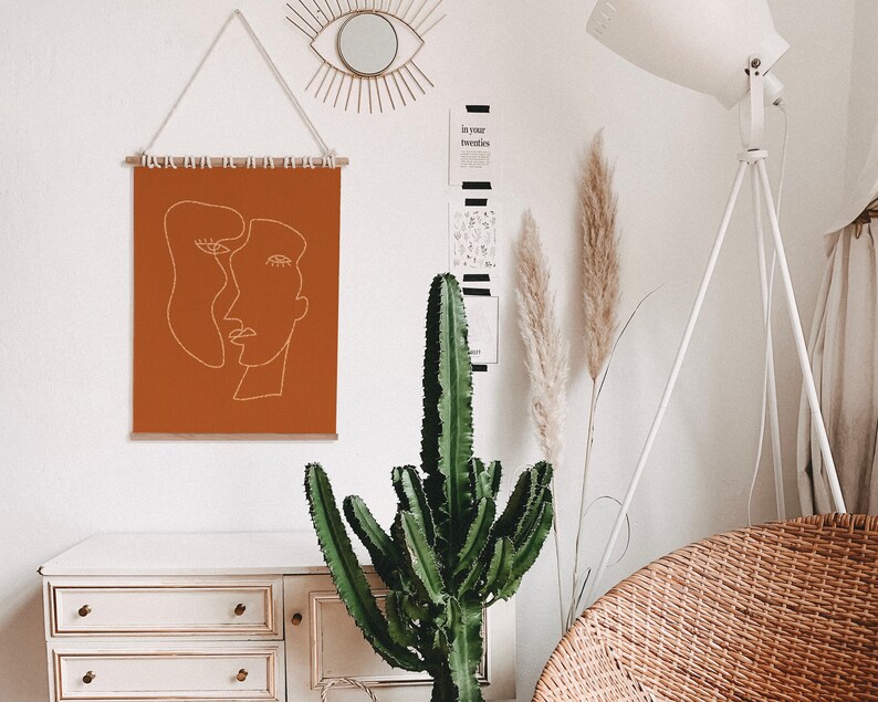 May include: A brown wall tapestry with a white line drawing of two abstract faces. The tapestry is hanging on a white wall with a decorative mirror above it. A large green cactus plant is in front of a white dresser with three drawers. A white floor lamp with a white shade is in the background.