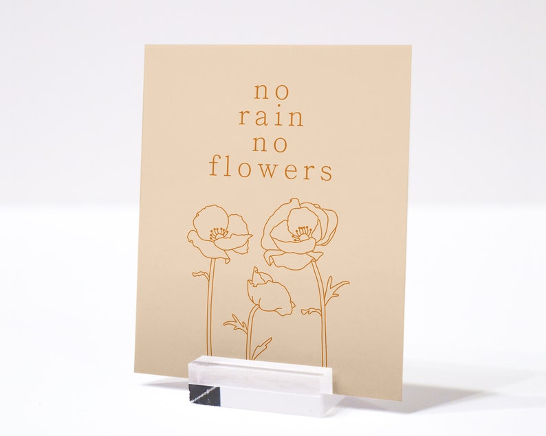 May include: A beige card with the text "no rain no flowers" in brown lettering. Three stylized poppy flowers are drawn in brown on the card.