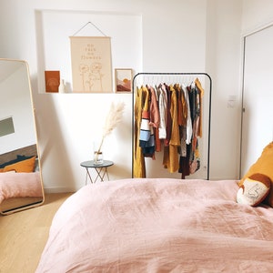May include: A bedroom with a pink duvet cover, a full-length mirror, a clothing rack with various garments, and a wall hanging with the text "no rain no flowers".
