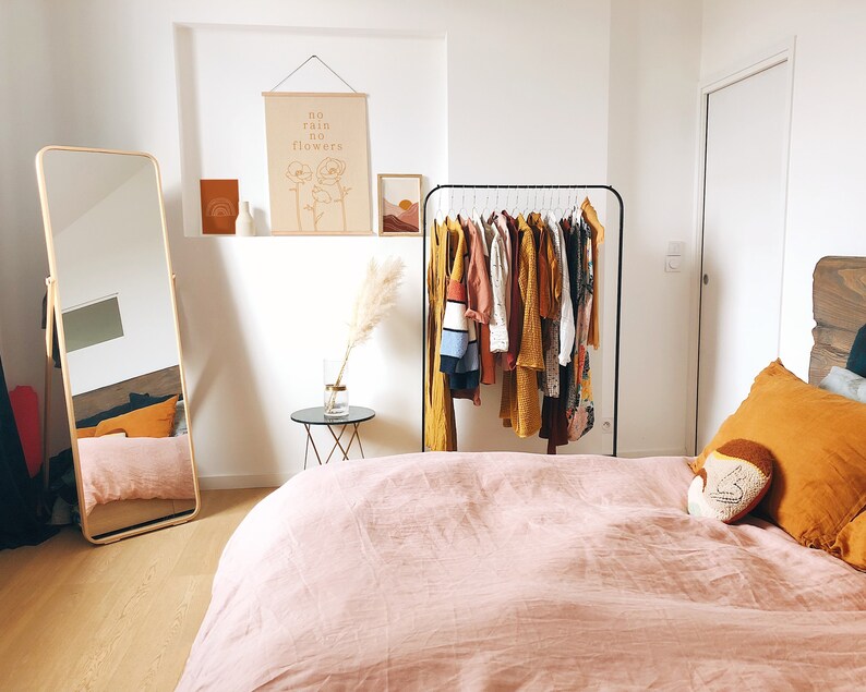 May include: A bedroom with a pink duvet cover, a full-length mirror, a clothing rack with various garments, and a wall hanging with the text "no rain no flowers".