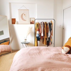 May include: A bedroom with a pink bedspread, a wooden headboard, and a clothing rack with a variety of clothes in shades of yellow, orange, and brown. A large mirror is on the left side of the room, and a small table with a vase of dried pampas grass is on the right side of the room.