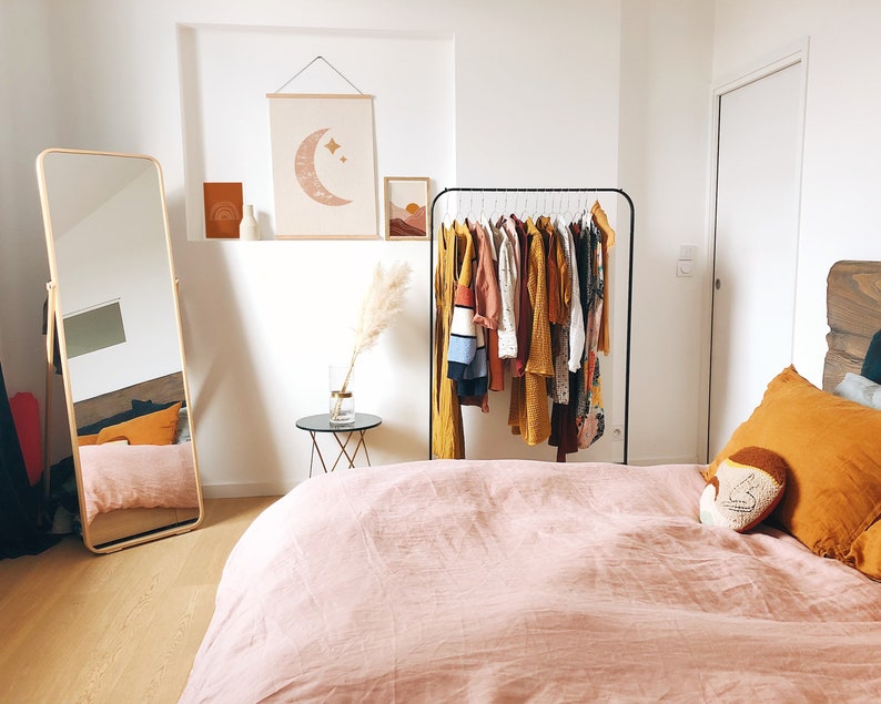 May include: A bedroom with a pink bedspread, a wooden headboard, and a clothing rack with a variety of clothes in shades of yellow, orange, and brown. A large mirror is on the left side of the room, and a small table with a vase of dried pampas grass is on the right side of the room.