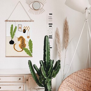 May include: A wall hanging featuring a stylized illustration of a leopard sitting in a jungle scene with green leaves and a black vase. The hanging is framed with a wooden dowel rod and has a white background.