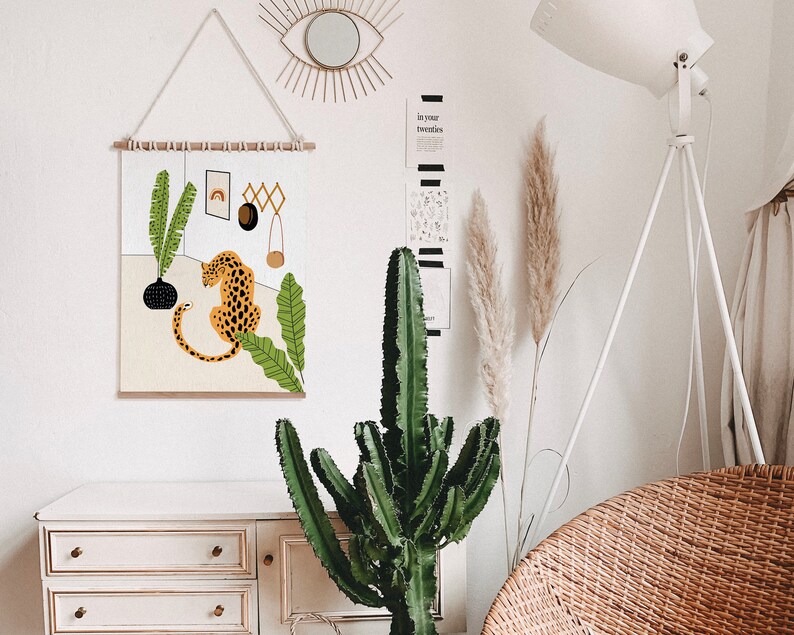 May include: A wall hanging featuring a stylized illustration of a leopard sitting in a jungle scene with green leaves and a black vase. The hanging is framed with a wooden dowel rod and has a white background.