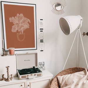 May include: A white record player with a brown and white record on a white shelf with a framed print of a potted plant on a brown background. The print is on a white wall with a white lamp with a white shade and a gold eye-shaped wall decor.