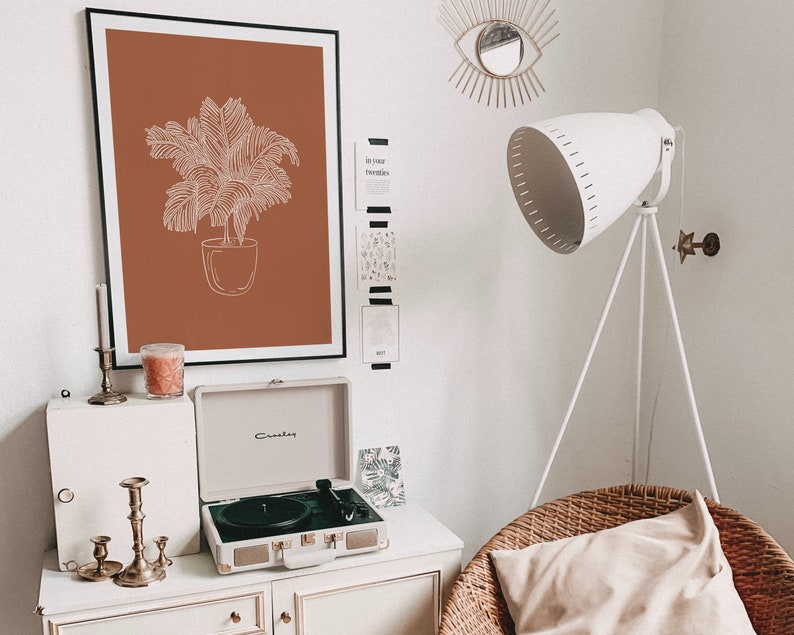 May include: A white record player with a brown and white record on a white shelf with a framed print of a potted plant on a brown background. The print is on a white wall with a white lamp with a white shade and a gold eye-shaped wall decor.