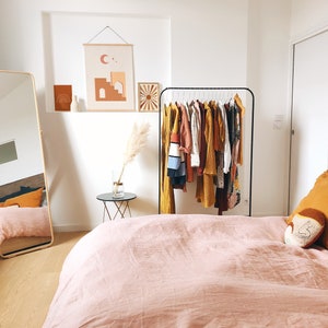May include: A bedroom with a pink linen duvet cover on the bed, a full-length mirror, a clothing rack with a variety of clothes, and a small side table with a vase of dried pampas grass.