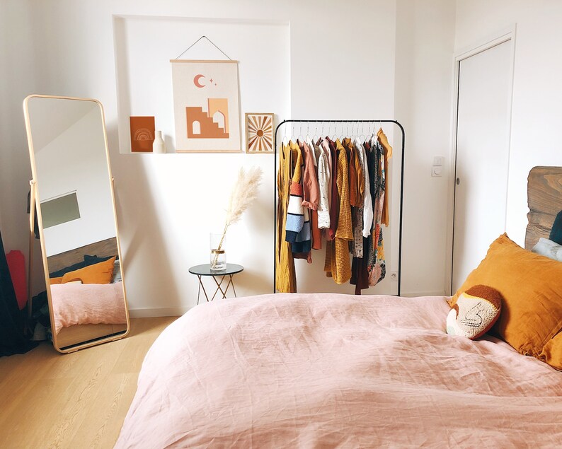 May include: A bedroom with a pink linen duvet cover on the bed, a full-length mirror, a clothing rack with a variety of clothes, and a small side table with a vase of dried pampas grass.
