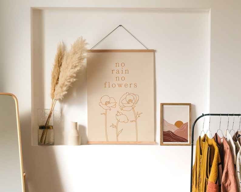 May include: A beige wall with a framed print that reads "no rain no flowers" with a line drawing of two flowers. There is a vase with dried pampas grass on a shelf below the print. A second framed print with a sunset landscape is to the right of the first print. A rack with clothes hangs on the right side of the image.