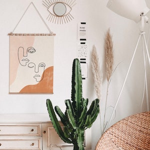 Puede incluir: Una pared blanca con una impresi&oacute;n de arte abstracto beige y naranja colgada. La impresi&oacute;n presenta tres caras con l&iacute;neas negras. Hay una l&aacute;mpara blanca con un tr&iacute;pode blanco, una gran planta de cactus verde y una c&oacute;moda blanca con tres cajones. La c&oacute;moda tiene pomos dorados. Hay dos plantas de hierba de la pampa secas en un jarr&oacute;n en la pared.