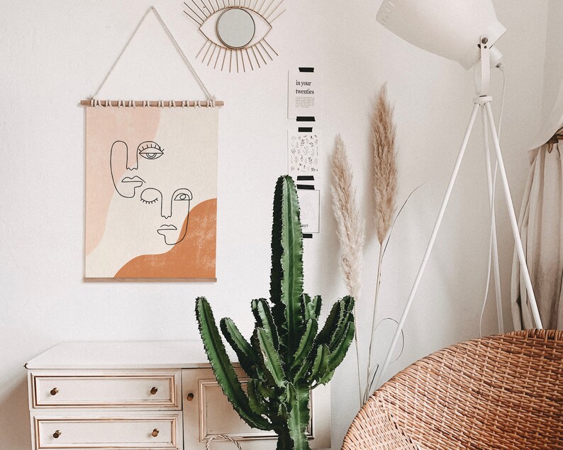 Puede incluir: Una pared blanca con una impresi&oacute;n de arte abstracto beige y naranja colgada. La impresi&oacute;n presenta tres caras con l&iacute;neas negras. Hay una l&aacute;mpara blanca con un tr&iacute;pode blanco, una gran planta de cactus verde y una c&oacute;moda blanca con tres cajones. La c&oacute;moda tiene pomos dorados. Hay dos plantas de hierba de la pampa secas en un jarr&oacute;n en la pared.