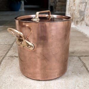 May include: A copper pot with a lid and brass handles. The pot is cylindrical with a shiny, reflective surface. The lid has a single handle, and the pot has two side handles. The pot is sitting on a stone surface.