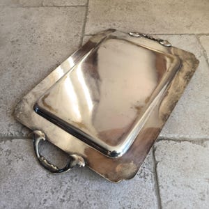 May include: A silver serving tray with two handles. The tray is rectangular and has a raised edge. The tray is made of polished silver and has a shiny finish.