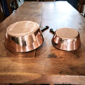 May include: Two copper saucepans with black handles sit on a wooden table. The pans have a shiny, reflective surface, highlighting their polished copper exterior. The handles are curved and appear to be made of a dark material.