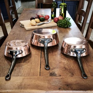 May include: Three copper saucepans with dark handles are displayed on a wooden table. The pans have a hammered finish and a shiny, reflective surface. A wooden cutting board with vegetables and olive oil bottles are in the background.
