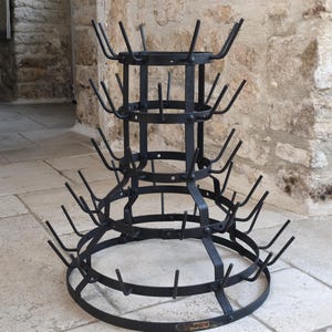Op de afbeelding: Een zwart metalen flessenrek met een gelaagde, conische structuur met vier cirkelvormige ringen van verschillende diameters. Elke ring is uitgerust met meerdere omhoog wijzende pinnen van donker ijzer voor het vasthouden van flessen.