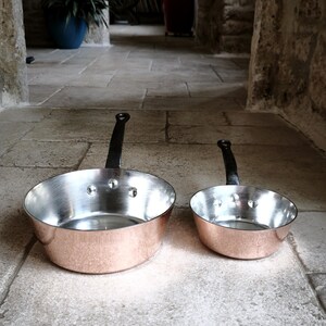 May include: Two copper-bottomed pans with silver interiors and black handles. The pans are of different sizes, with the larger one on the left and the smaller one on the right. The pans are on a stone surface.