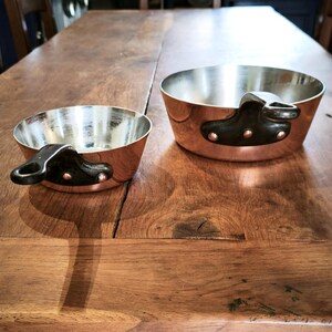 May include: Two copper saucepans with silver interiors and black handles. The pans are different sizes, with the larger one having a diameter of approximately 20 cm. The handles are attached with rivets.