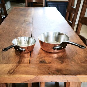 May include: Two copper saucepans with black handles on a wooden table. The pans have a shiny interior and a rounded shape. The handles are riveted to the pans. One pan is smaller than the other.