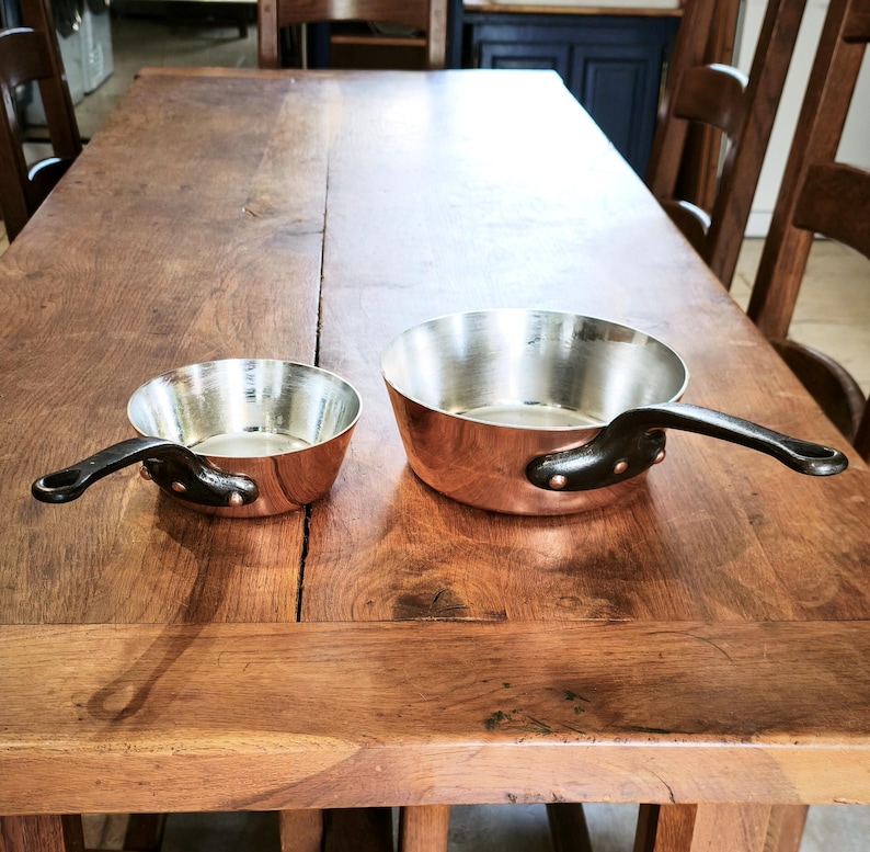 May include: Two copper saucepans with black handles on a wooden table. The pans have a shiny interior and a rounded shape. The handles are riveted to the pans. One pan is smaller than the other.