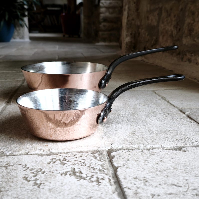 May include: Two copper pans with black handles are shown on a stone surface. The pans have a shiny, silver interior and a hammered copper exterior. The handles are curved and attached with rivets. These pans are likely for cooking.