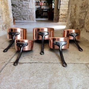 May include: Five copper saucepans with dark metal handles are arranged on a stone surface. The pans have a shiny, reflective surface and a classic design. The handles are riveted to the pans.