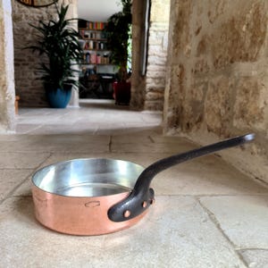 May include: A copper saucepan with a silver interior and a long, black handle. The pan is round with a slightly flared rim. The handle is attached with rivets. The pan is on a stone floor.