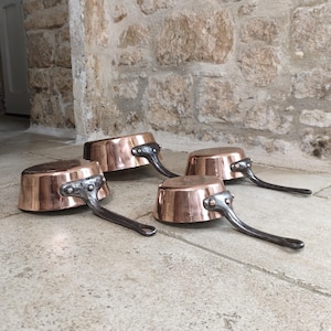 Puede incluir: Cuatro cacerolas de cobre con asas de metal oscuro dispuestas sobre una superficie de piedra. Las cacerolas tienen una superficie brillante y reflectante y un diseño clásico. Las asas están remachadas a los cuerpos de cobre.