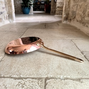 May include: A copper-colored saucepan with a long, brass-colored handle. The pan has a rounded shape and a polished, reflective surface. The handle is attached to the pan with rivets. The cookware is designed for cooking.