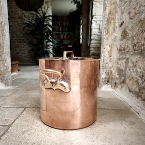 May include: A cylindrical copper container with a lid and decorative handles. The container has a polished finish and is set on a stone surface. The handles and lid handle are a contrasting gold colour. The container is likely for kitchen or home decor.