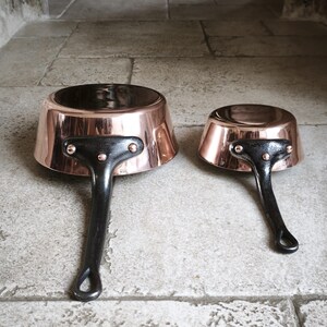 May include: Two copper saucepans with black handles. The pans have a shiny copper exterior and a dark interior. The handles are riveted to the pans. The pans are of different sizes, with the larger one measuring approximately 18 cm in diameter.