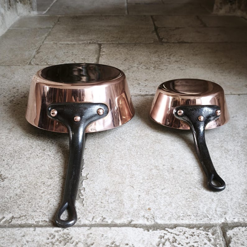 May include: Two copper saucepans with black handles. The pans have a shiny copper exterior and a dark interior. The handles are riveted to the pans. The pans are of different sizes, with the larger one measuring approximately 18 cm in diameter.
