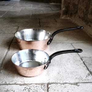 May include: Two copper saucepans with black handles. The interior of the pans is silver. The pans are on a stone surface. The handles are curved and attached to the side of the pans.