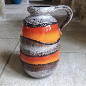 Może przedstawiać: Ceramiczny dzbanek z uchwytem, ​​z poziomymi pasami w kolorze pomarańczowym, czarnym i srebrnym. Dzbanek ma teksturowaną powierzchnię i zaokrągloną podstawę. Uchwyt zakrzywia się od góry dzbanka.