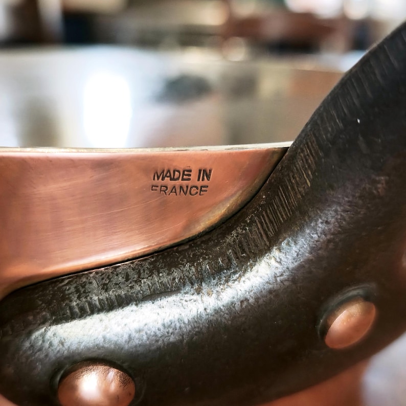 May include: Close-up of a copper cooking pot with a dark handle. The pot has a shiny, reflective surface. The text "MADE IN FRANCE" is stamped on the side of the pot.