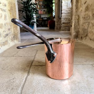 May include: A copper saucepan with a dark metal handle and rivets. The pot has a shiny, reflective surface and is cylindrical in shape. The handle curves upwards and away from the pot's body. The pot is sitting on a stone surface.