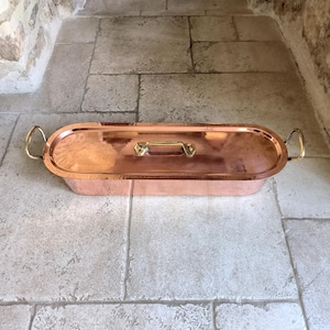Puede incluir: Una fuente para asar ovalada de color cobre con tapa. La fuente tiene asas de lat&oacute;n en cada extremo y una asa de lat&oacute;n en la tapa. La fuente est&aacute; sobre una superficie de piedra.