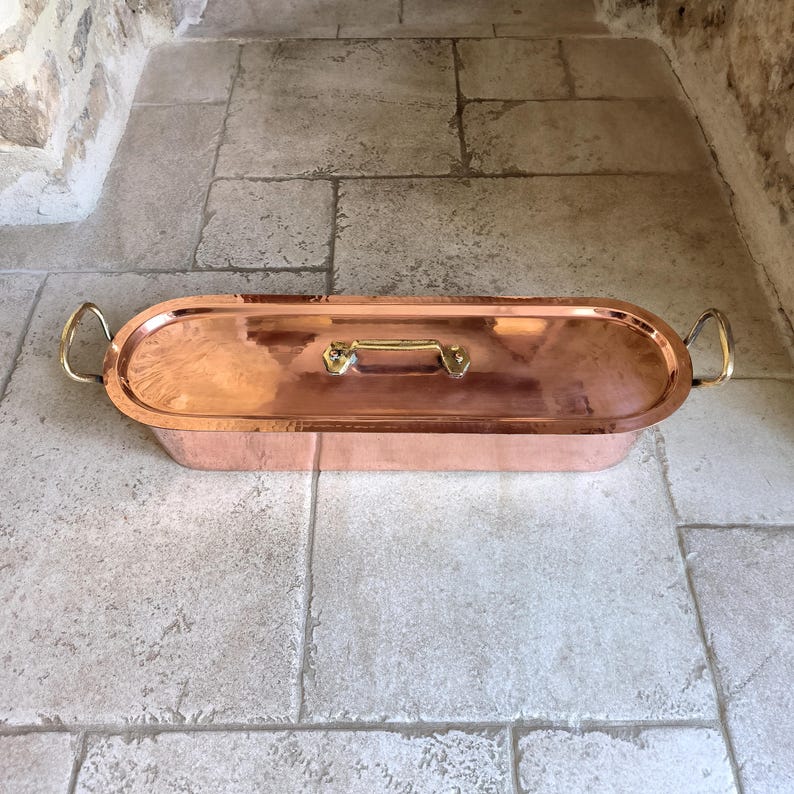 Puede incluir: Una fuente para asar ovalada de color cobre con tapa. La fuente tiene asas de lat&oacute;n en cada extremo y una asa de lat&oacute;n en la tapa. La fuente est&aacute; sobre una superficie de piedra.