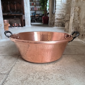 May include: A large copper bowl with a hammered finish and two black metal handles. The bowl is sitting on a tiled floor.