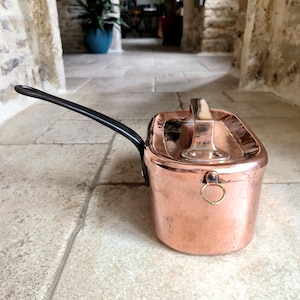 May include: A copper saucepan with a black handle and a copper lid. The saucepan has a rectangular shape and a small brass ring attached to the side. The handle is curved and attached to the side of the pan.