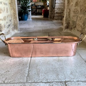 Puede incluir: Una asadera rectangular de cobre con asas y tapa de lat&oacute;n. La sart&eacute;n tiene un acabado pulido y est&aacute; sobre una superficie de piedra. Las asas son curvas y est&aacute;n unidas a los lados de la sart&eacute;n.