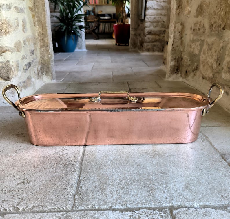 Puede incluir: Una asadera rectangular de cobre con asas y tapa de lat&oacute;n. La sart&eacute;n tiene un acabado pulido y est&aacute; sobre una superficie de piedra. Las asas son curvas y est&aacute;n unidas a los lados de la sart&eacute;n.