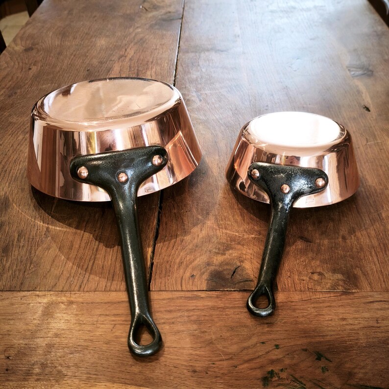 May include: Two copper saucepans with dark green handles. The pans have a shiny copper exterior and a lighter interior. The handles are riveted to the pans and have a loop at the end. The pans are on a wooden surface.