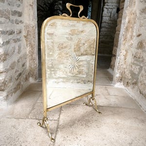 May include: A vintage brass vanity mirror with a rectangular frame and a decorative starburst design etched into the glass. The mirror has a curved handle and ornate, scrolled feet. The brass has a warm, aged patina.
