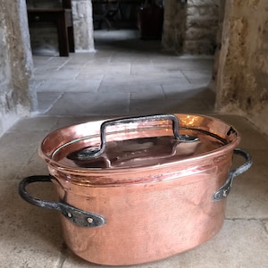 Può includere: Una pentola ovale in rame con due manici in ferro nero e un manico per il coperchio in ferro nero. La pentola ha una finitura martellata e si trova su un pavimento piastrellato.