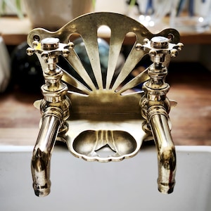 Op de afbeelding: Close-up van een vintage messing kraan met twee tuiten. De kraan heeft een decoratieve achterplaat met een sunburst design. De handvatten zijn kruisvormig. De kraan is gemonteerd op een witte wastafel.